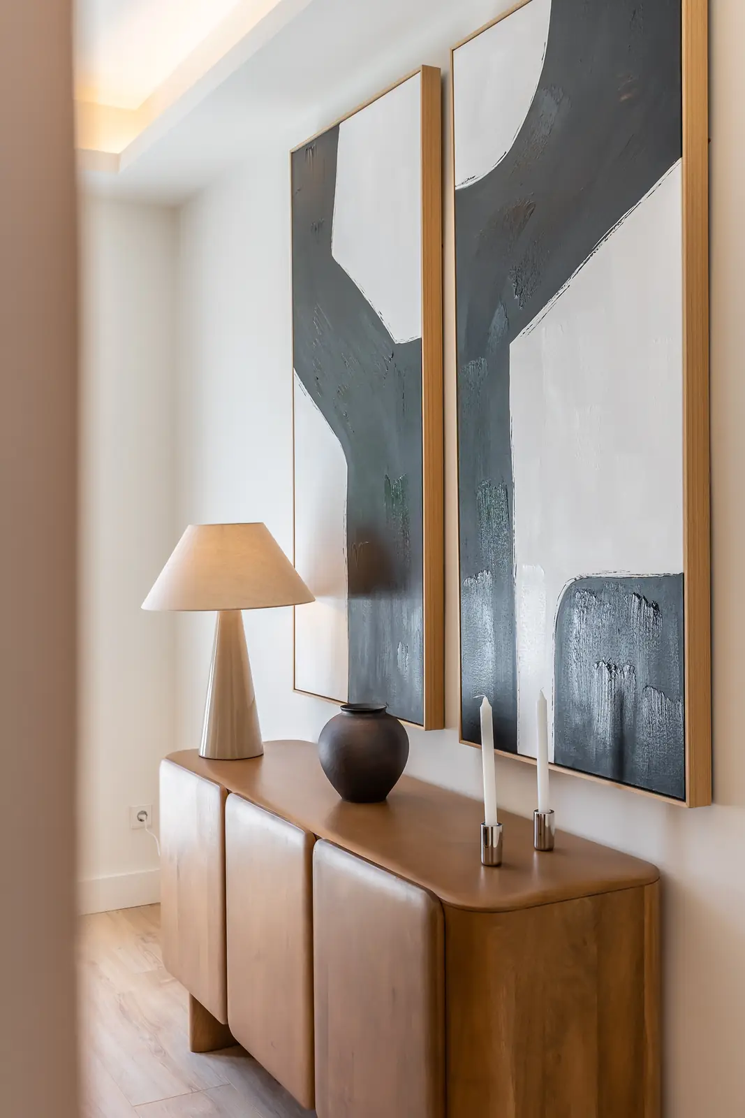 Recibidor de salón con aparador de madera curvada, lámpara y cuadros abstractos